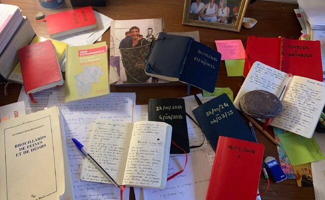 Photographie de la table de travail du compagnon d'Émilie Aussel (carnets, photos, stylos...)