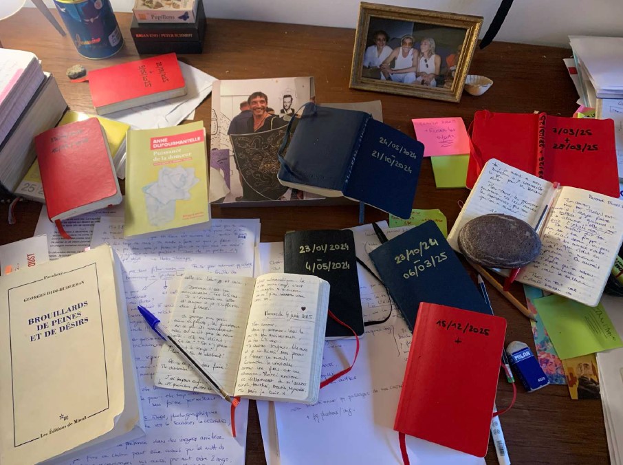 Photographie de la table de travail du compagnon d'Émilie Aussel (carnets, photos, stylos...)